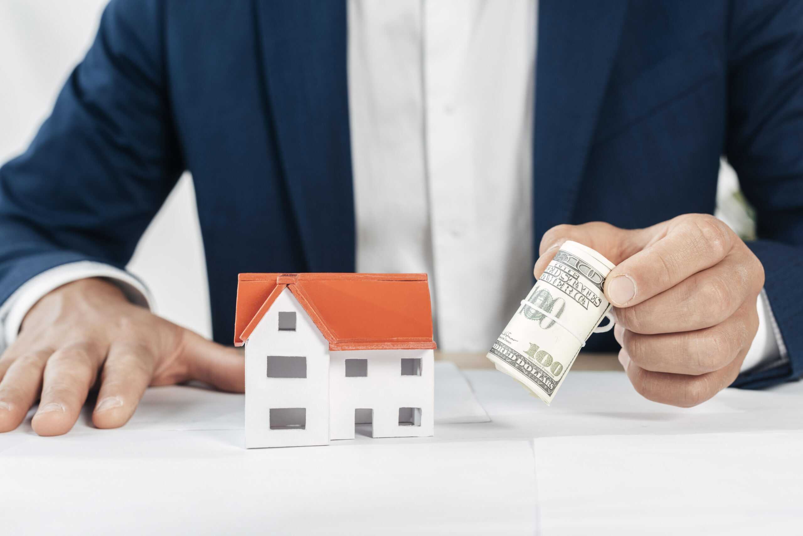 Hand holding cash near a small house model for home sale